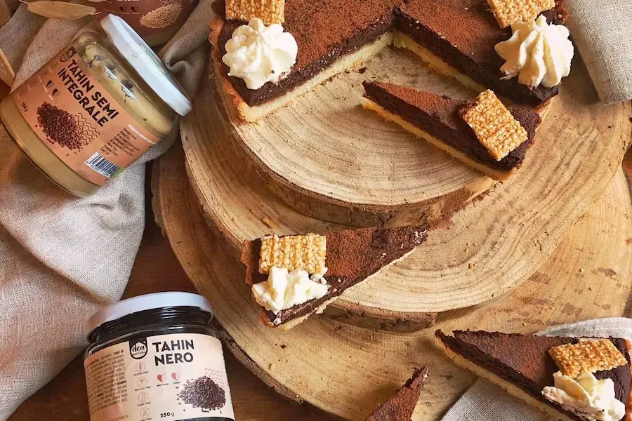 Crostata tahin e cioccolato su un tagliere in legno e dietro 3 vasetti diversi di Tahin