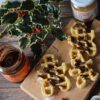 Tavolo di di legno con sopra agrifoglio di natale, 2 vasetti di creme spalmabili una al gusto di pure pasta di arachidi 100% e l altro senza tappo al gusto di mandorla e cacao. Inoltre in primo piano troviamo un vassoio di legno con dei waffle realizzati con pasta pura di arachidi 100% a forma di omino di pan di zenzero con sopra crema mandorla e cacao.