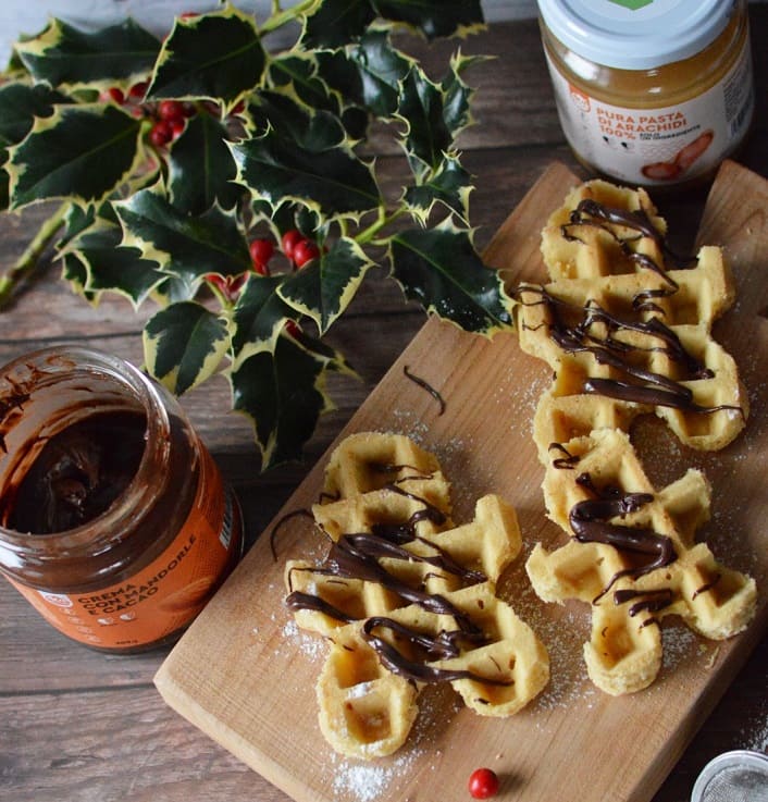 Tavolo di di legno con sopra agrifoglio di natale, 2 vasetti di creme spalmabili una al gusto di pure pasta di arachidi 100% e l altro senza tappo al gusto di mandorla e cacao. Inoltre in primo piano troviamo un vassoio di legno con dei waffle realizzati con pasta pura di arachidi 100% a forma di omino di pan di zenzero con sopra crema mandorla e cacao.