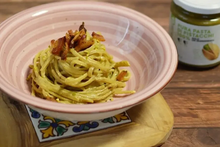 Su di un tavolo di legno troviamo un piatto fondo con delle linguine con crema di pistacchio 100% dea nocciola e pancetta croccante. Inoltre a destra del piatto troviamo un vasetto di pura pasti di pistacchi 100% deanocciola
