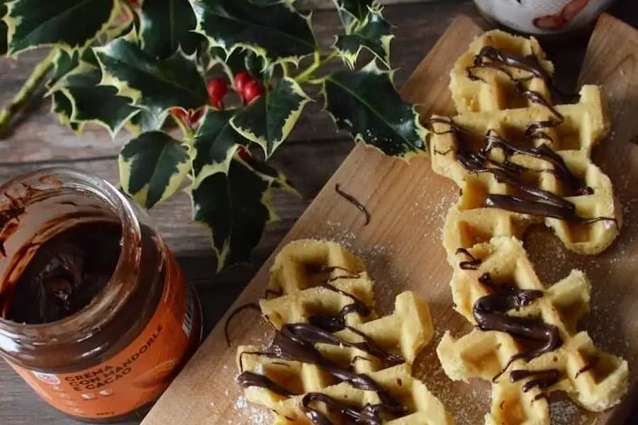 Tavolo di di legno con sopra agrifoglio di natale, 2 vasetti di creme spalmabili una al gusto di pure pasta di arachidi 100% e l altro senza tappo al gusto di mandorla e cacao. Inoltre in primo piano troviamo un vassoio di legno con dei waffle realizzati con pasta pura di arachidi 100% a forma di omino di pan di zenzero con sopra crema mandorla e cacao.