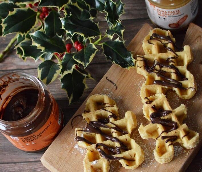 Tavolo di di legno con sopra agrifoglio di natale, 2 vasetti di creme spalmabili una al gusto di pure pasta di arachidi 100% e l altro senza tappo al gusto di mandorla e cacao. Inoltre in primo piano troviamo un vassoio di legno con dei waffle realizzati con pasta pura di arachidi 100% a forma di omino di pan di zenzero con sopra crema mandorla e cacao.