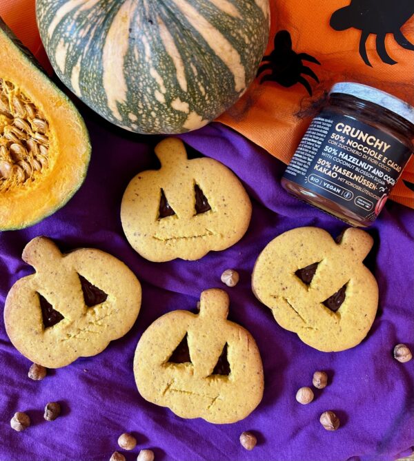 Tovaglia viola e arancio, con adagiati biscotti a forma di zucca ripieni di crema crunchy 50% nocciole e cacao, 2 zucche di cui una aperta a metà e un vasetto di crema spalmabile al gusto Crunchy 50% nocciole e cacao.