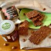 Barrette snack senza glutine adagiate su di un piatto verde e una tovaglietta beige. Nella parte a destra troviamo un vasetto 3 ingredienti Nocciola,Zucchero di cocco e Cacao e un vasetto di proteica vegan Nocciola e Cacao
