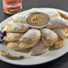 Ravioli dolci ripieni di crema al gusto cappuccino su un piatto bianco, ricoperti da zucchero a velo. Sopra ai ravioli, nella parte centrale troviamo un piattino con all interno la crema al cappuccino. Nella parte alta a sinistra dello sfondo troviamo il vasetto di crema al gusto cappuccino.
