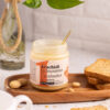 tavola beige con barattolo di crema 100% arachidi aperto con cucchiaino dorato su una vassoio di legno, a destra tre fette biscottate e arachidi sparse. vaso con piante sullo sfondo