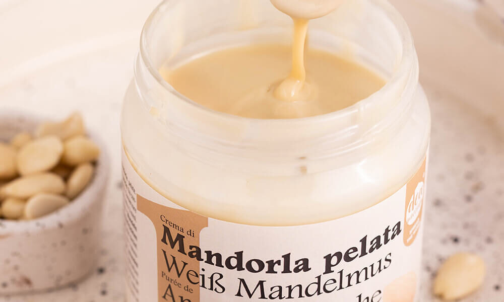 close up di crema 100% di mandorla pelata aperta con cucchiaino che cola sopra, il barattolo è dentro un vassoio bianco e ciotolina di mandorle a sinistra, mandorle pelate intorno