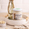crema 100% di mandorla pelata dentro un vassoio bianco e bottiglia con piante. a sinistra ciotolina di mandorle e mandorle pelate intorno