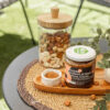 composizione con crema tre ingredienti con nocciola, ciotolina con zucchero, nocciole sparse, barattolo con frutta secca su un vassoio di legno e tovaglietta di raffia, su uno tavolino in un giardino