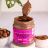 close up di un barattolo di crema crunchy nocciola e cacao aperto con sopra un cucchiaino colmo di crema. il barattolo è su una base effetto granito con ciotola di nocciole a sinistra e di cacao a destra, foglia sullo sfondo