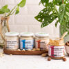 gruppo di creme 100% frutta secca dentro un vassoio di legno su una tavola effetto legno. fritta secca sparsa intorno. Vasi con piante sullo sfondo a destra e sinistra. Crema 100% nocciola fuori dal vassoio con cucchiaino dentro