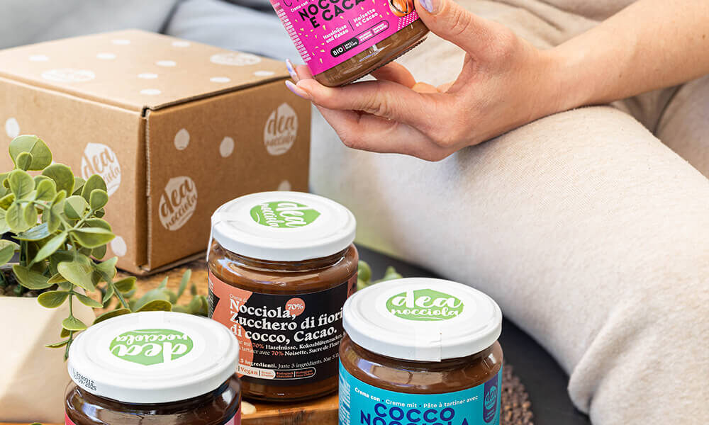 creme spalmabili su un tavolino, fuori e dentro un vassoio di legno su tovaglietta di raffia. A destra zoom su una ragazza seduta vestita di beige che legge una crema. scatola spedizioni deanocciola sullo sfondo, piantine finte a sinistra della composizione