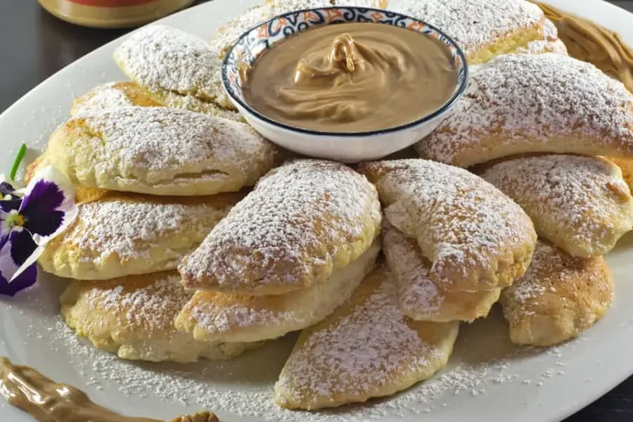 Ravioli dolci ripieni di crema al gusto cappuccino su un piatto bianco, ricoperti da zucchero a velo. Sopra ai ravioli, nella parte centrale troviamo un piattino con all interno la crema al cappuccino. Nella parte alta a sinistra dello sfondo troviamo il vasetto di crema al gusto cappuccino.