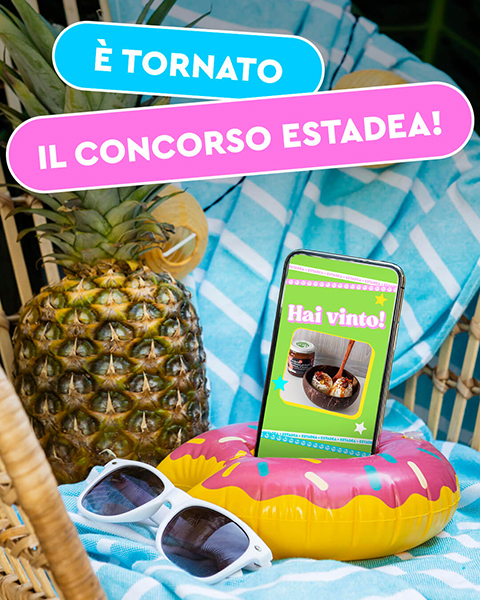 smartphone con fotografia vincitrice in una ciambella gonfiabile. sfondo con ananas, telo mare azzurro e occhiali da sole. testo: è tornato il concorso estadea